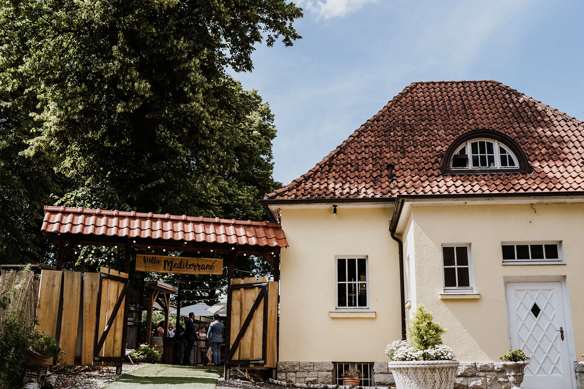 Eingang der Eventlocation Villa Mediterrané mit Holztor und rotem Ziegeldach, umgeben von Bäumen